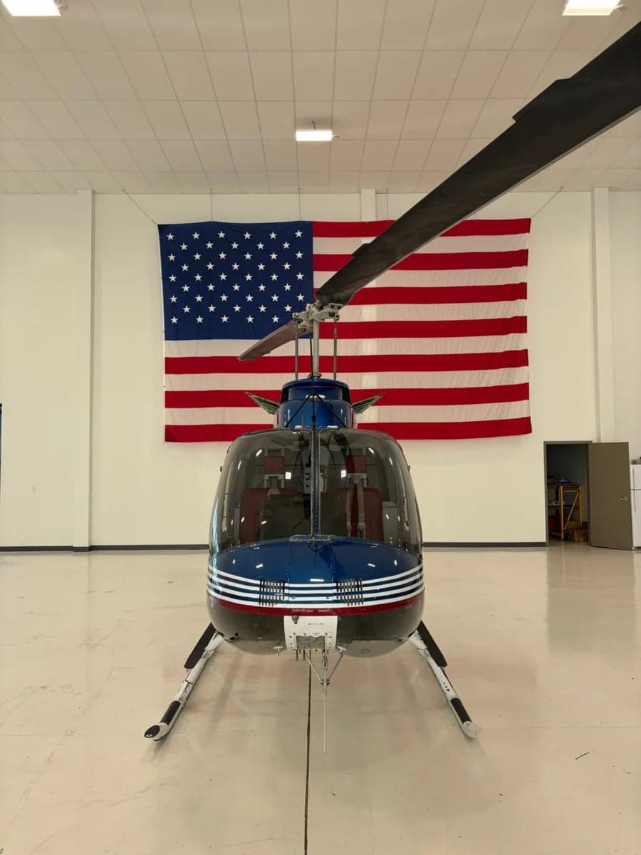 Front view of a blue and white Bell 206B JetRanger helicopter parked in a hangar with an American flag in the background.
