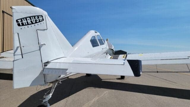 Rear-right view of a white Thrush agricultural aircraft with a black stripe on the fuselage and "THRUSH AIRCRAFT" on the tail.