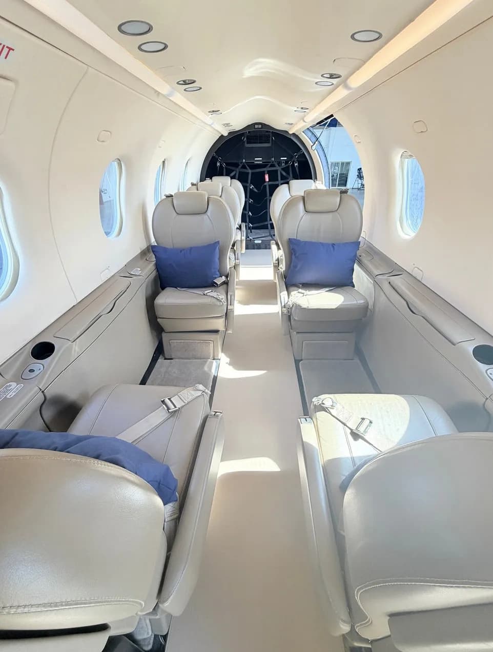 The interior cabin of a Pilatus PC-12 aircraft, featuring tan leather seats with blue accent pillows arranged in a club configuration.