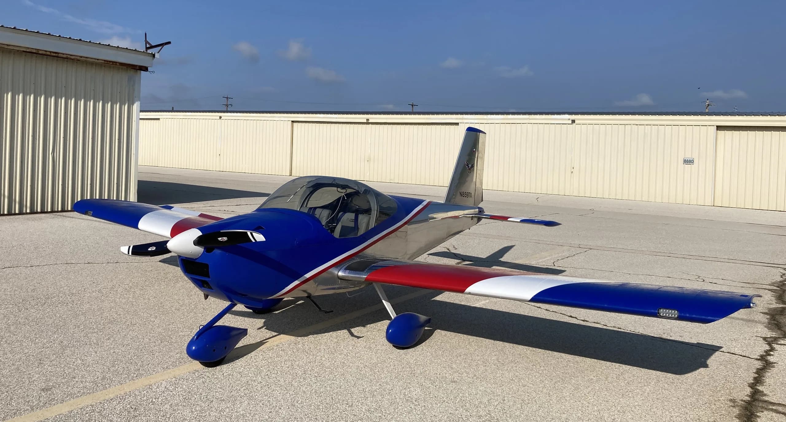 A blue, silver, red, and white Van's RV-7 aircraft is parked on an airfield ramp next to hangars under a clear sky.