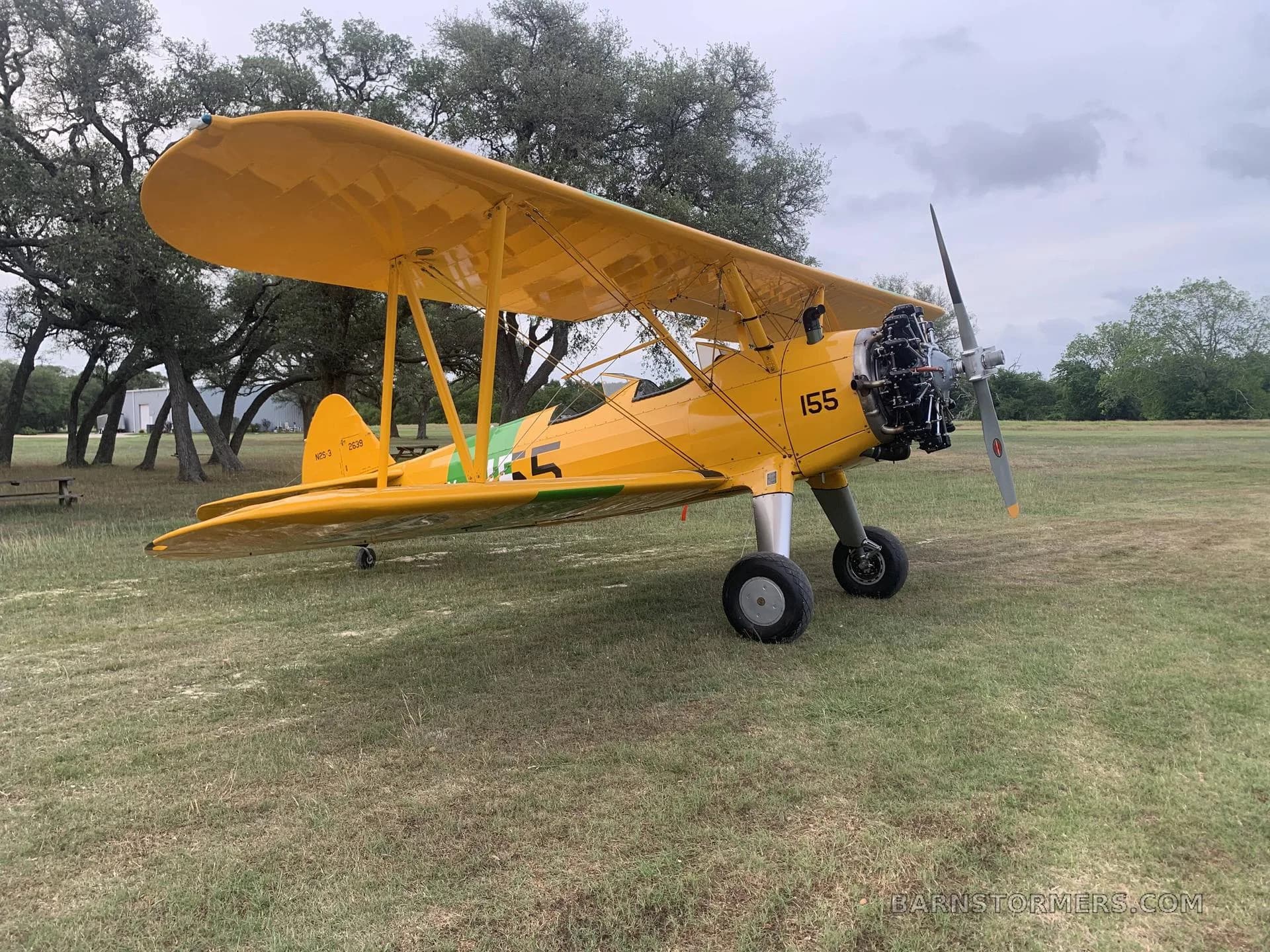 A yellow biplane with U.S. Navy markings and the number 155, parked on a grassy field with trees in the background.