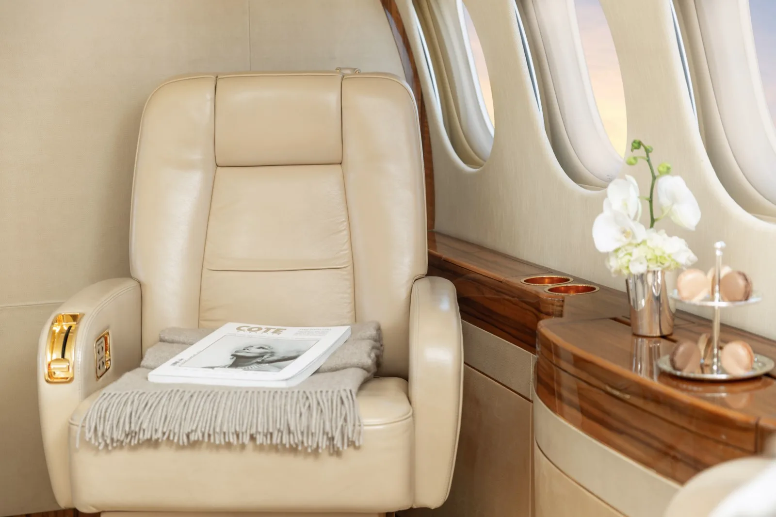 A cream leather passenger seat with a magazine and blanket, next to a wooden side table with flowers and macarons.