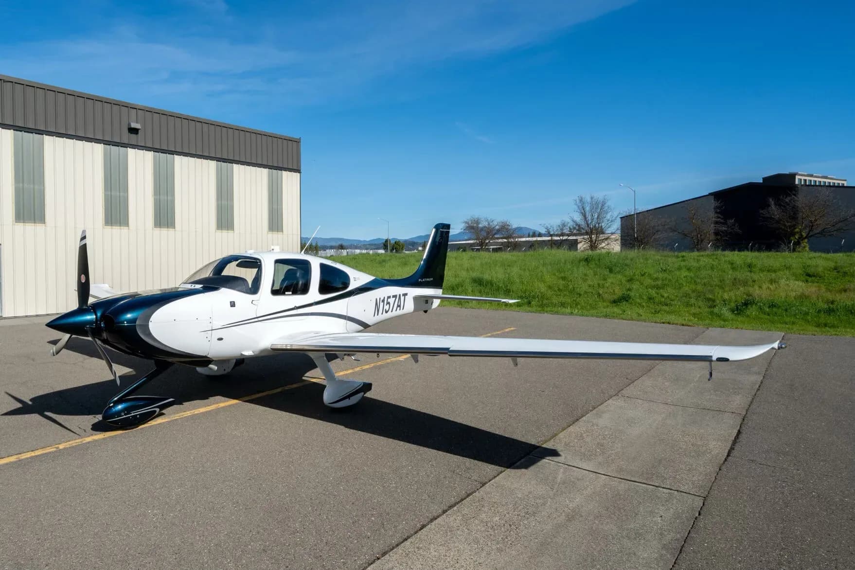 A white and dark blue Cirrus SR22 aircraft, registration N157AT, parked on an asphalt apron next to a hangar.