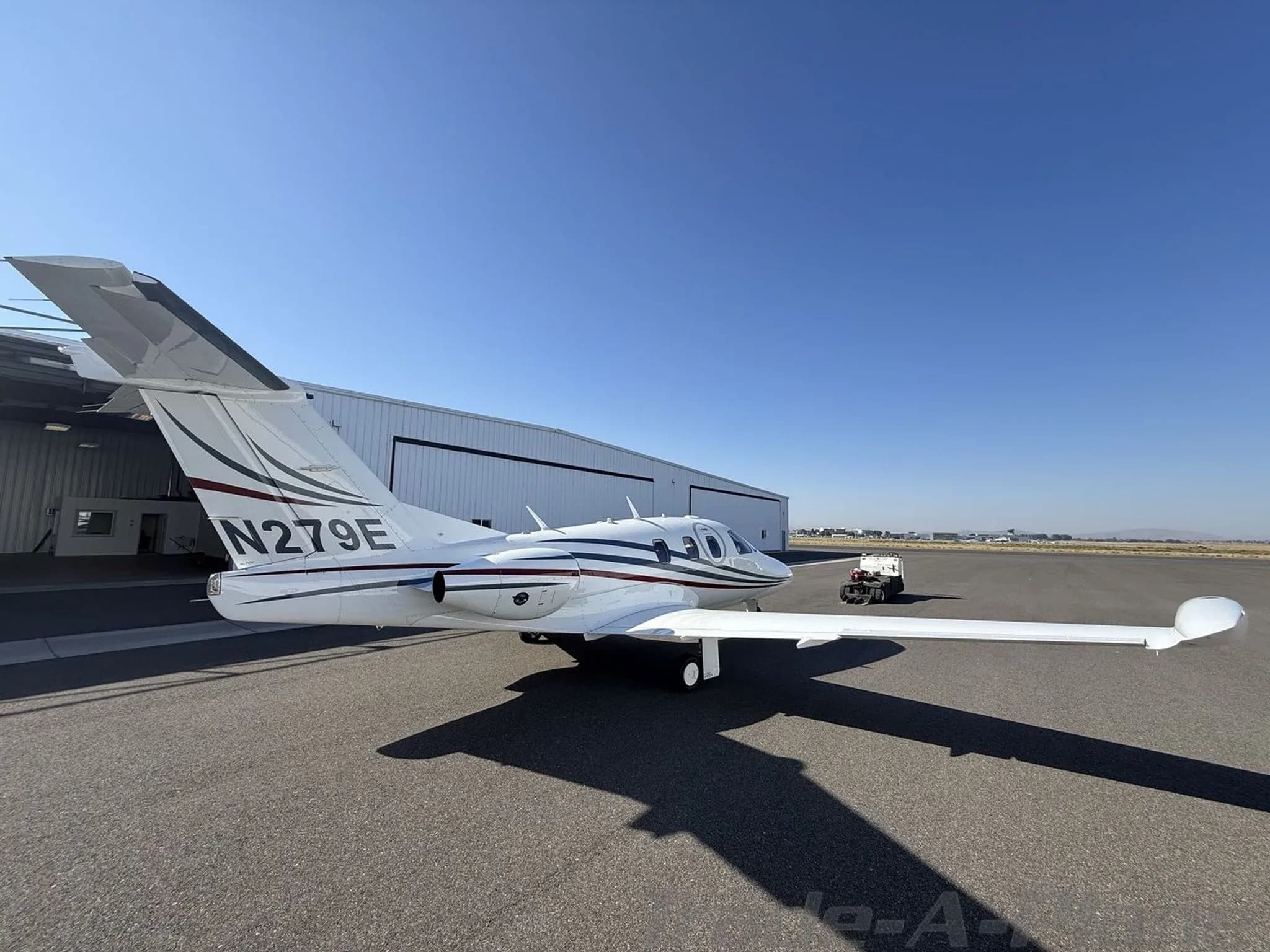 Rear-side view of a white private jet with red and blue stripes and registration N279E parked on an apron.