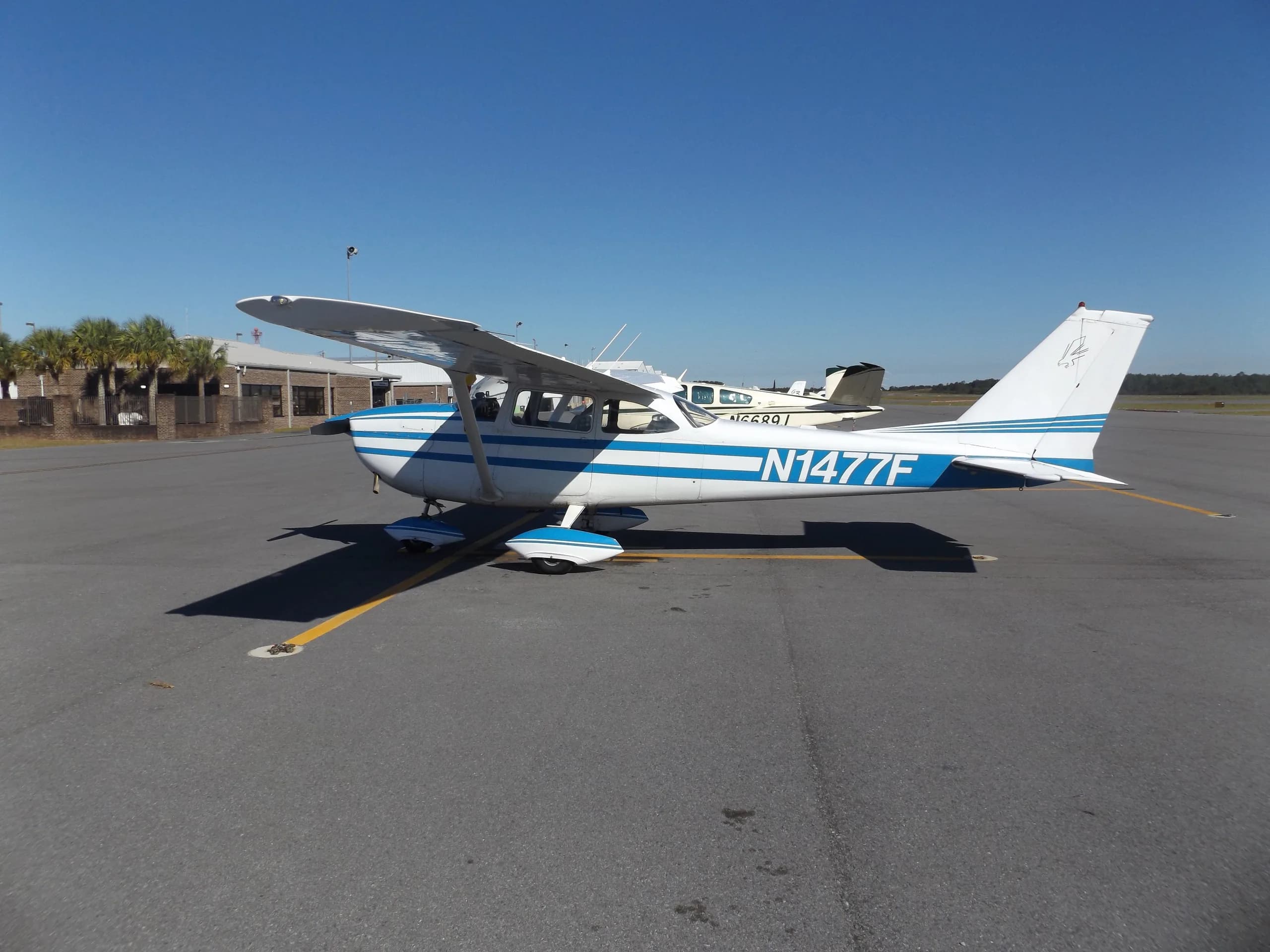 A white single-engine propeller aircraft with blue stripes (N1477F) parked on an airport tarmac.