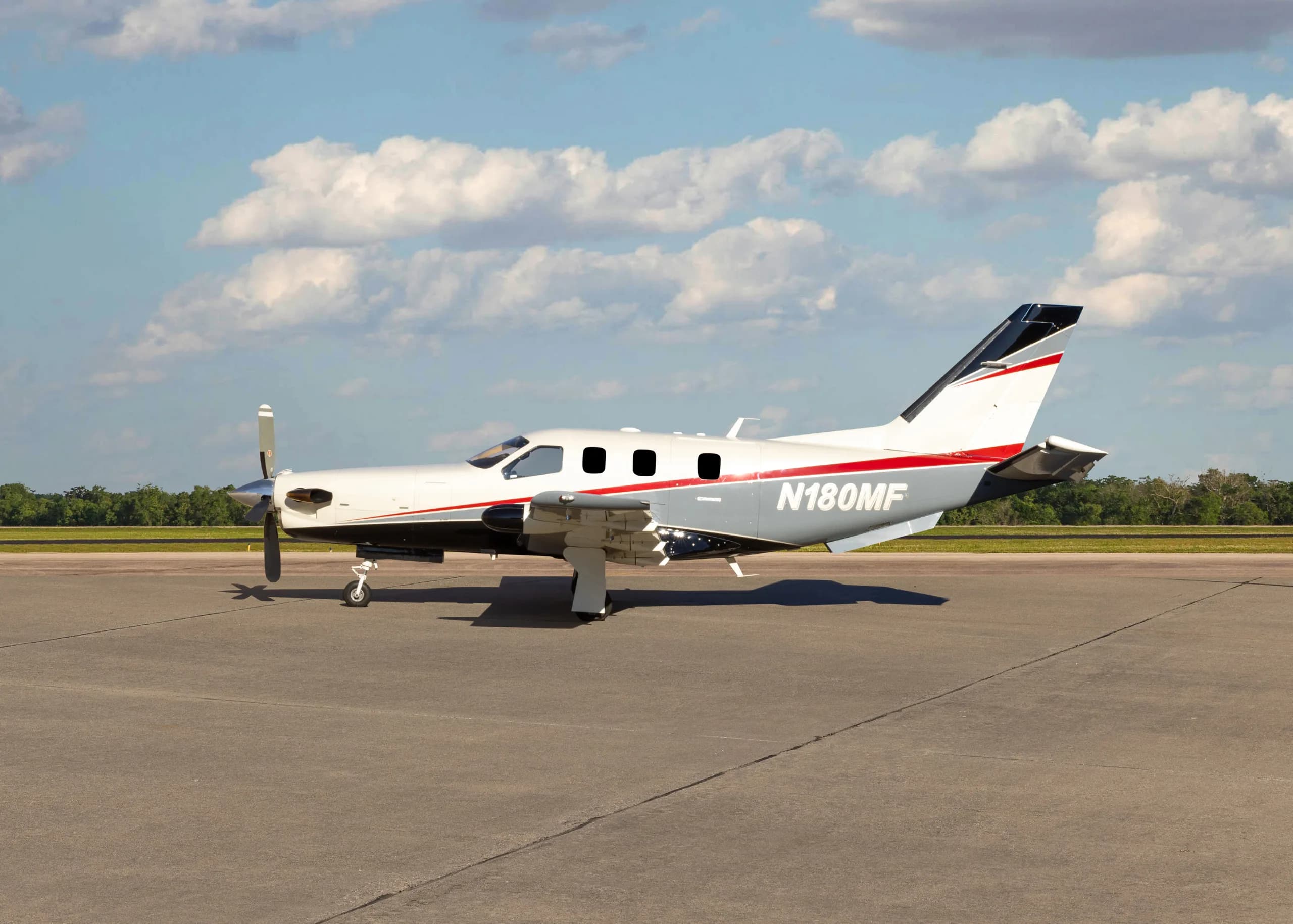 A white and grey TBM 900 series aircraft with red stripes, registration N180MF, parked on an airfield, showing the full left side.