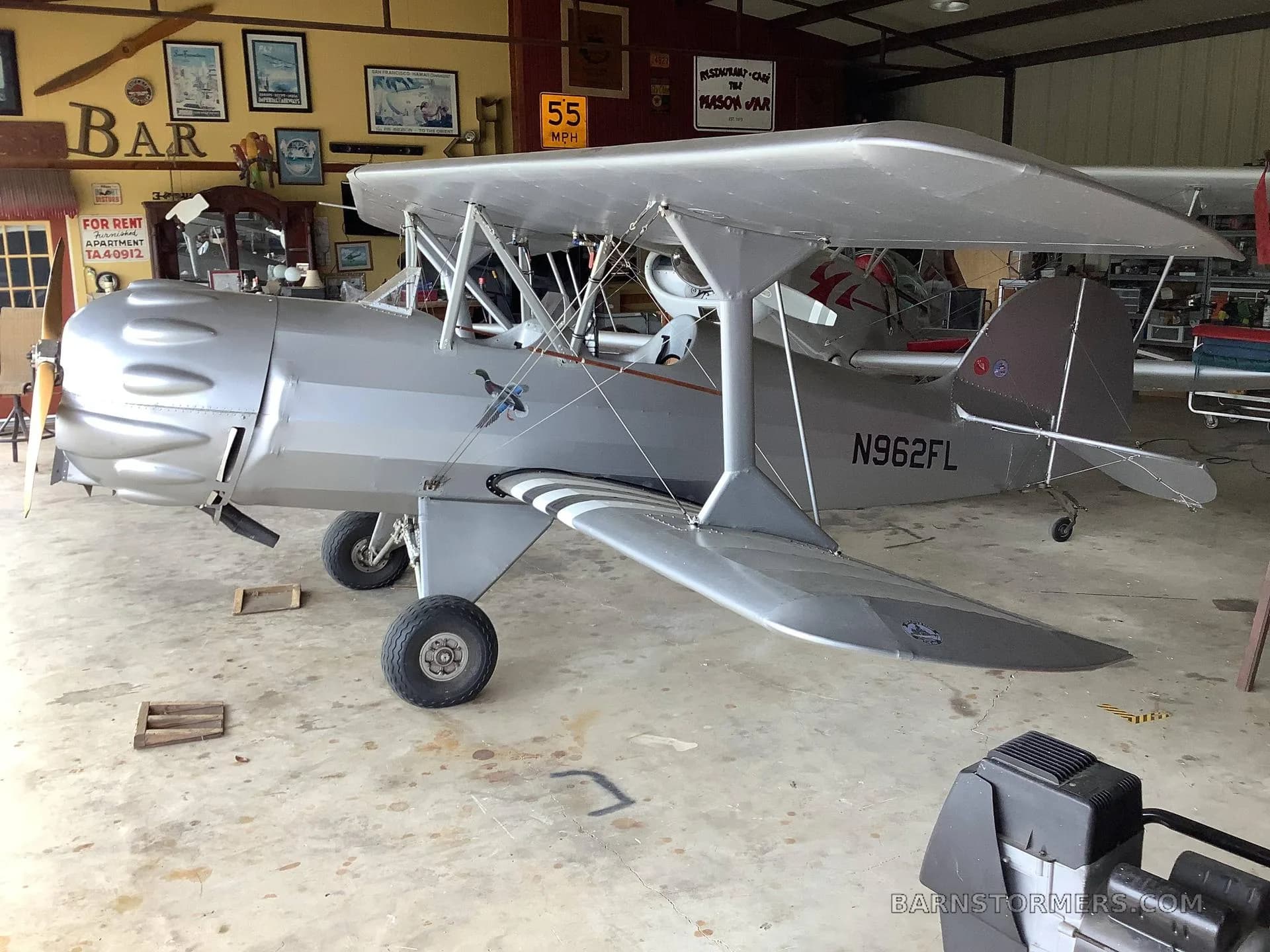A silver biplane with tail number N962FL is parked in a hangar, showing its side profile.