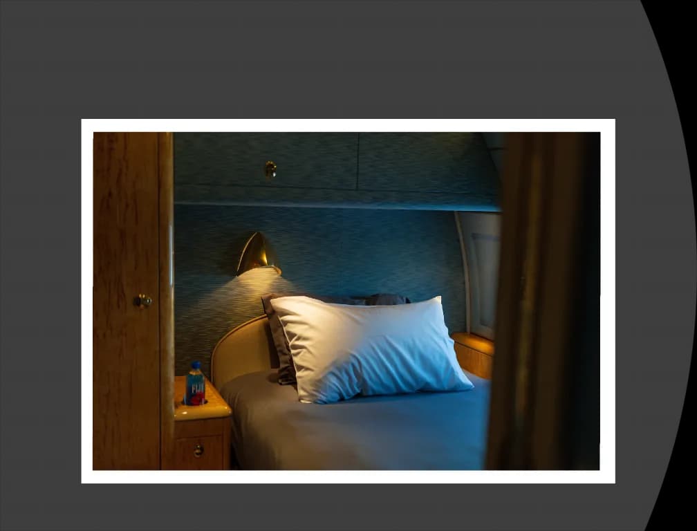 A private jet bedroom showing a single pillow on a bed with a bedside table and a bottle of Fiji water.