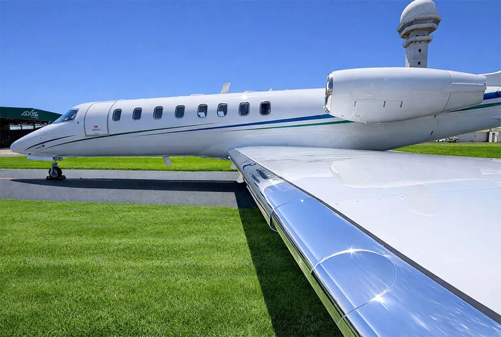 A close-up side view of a white business jet with blue and green stripes, highlighting the reflective wing surface and an airport control tower in the background.