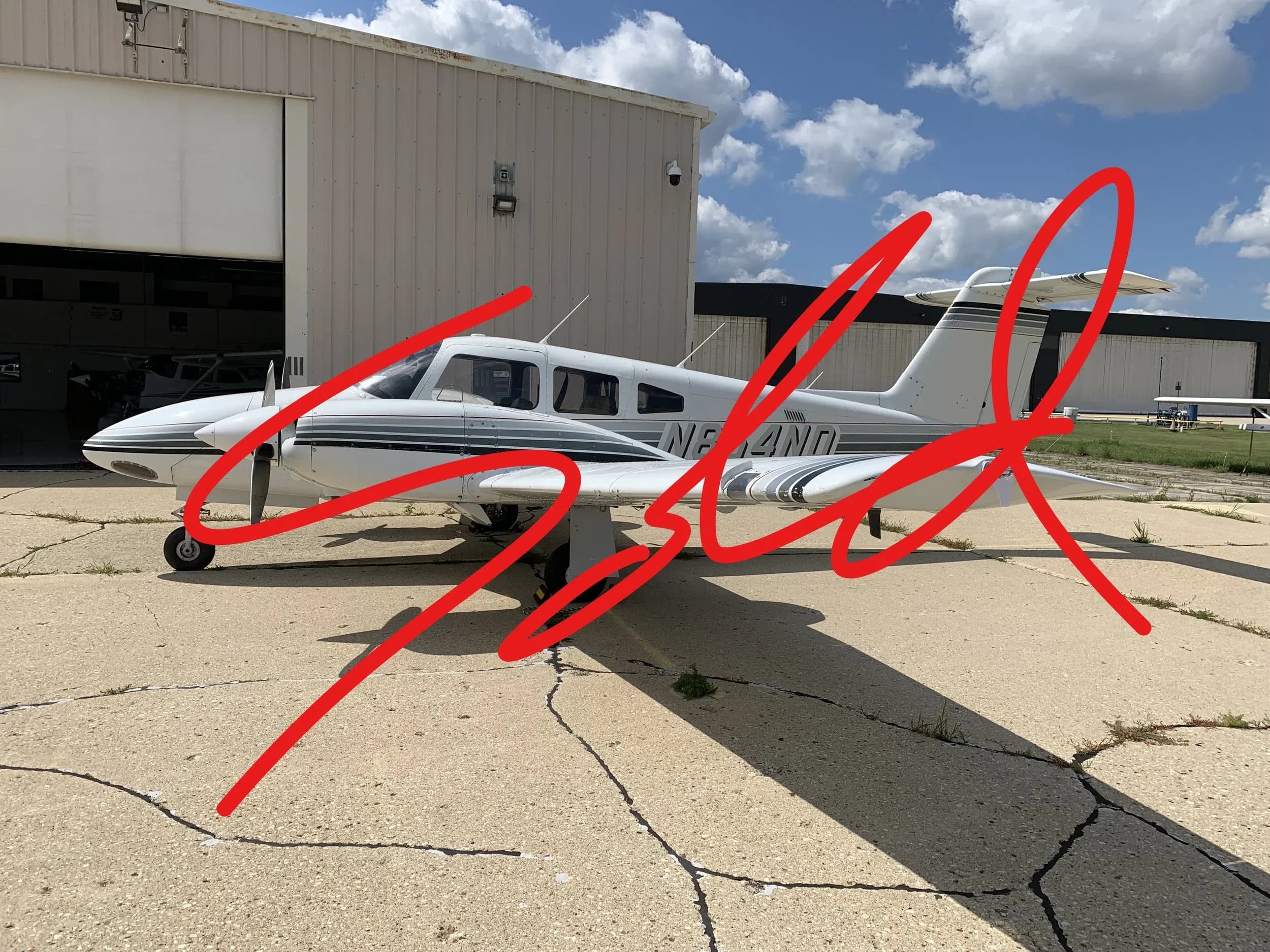 A white Piper Saratoga aircraft with grey stripes parked on a tarmac next to a hangar under a partly cloudy sky.