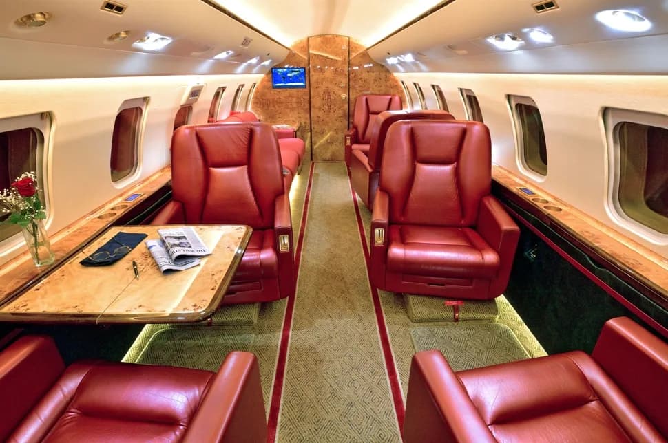 A luxurious aircraft cabin interior featuring red leather seats, light wood tables, and a TV screen.