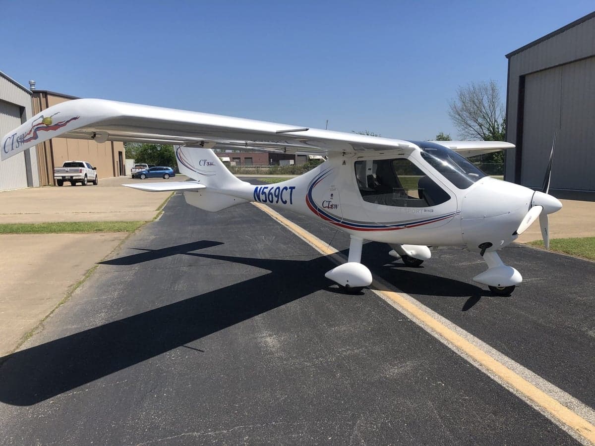 A white Flight Design CTsw light sport aircraft with red, blue, and grey stripes is parked on an asphalt ramp on a sunny day.