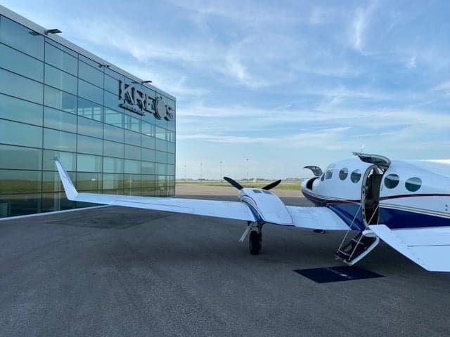 A white and blue twin-engine aircraft is parked on the tarmac with the door open.