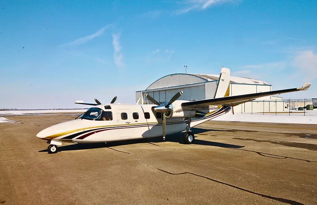 A white twin-engine turboprop aircraft with gold and maroon stripes parked on an airfield with a hangar in the background.
