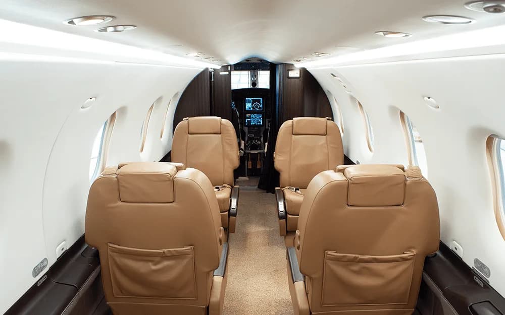 The aircraft cabin interior features two rows of tan leather seats facing forward, with the cockpit visible in the distance.