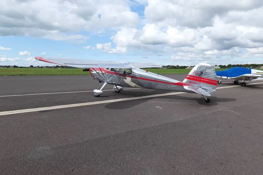 A side view of a silver Cessna 170B with red accents, parked on an airfield with another aircraft in the background.