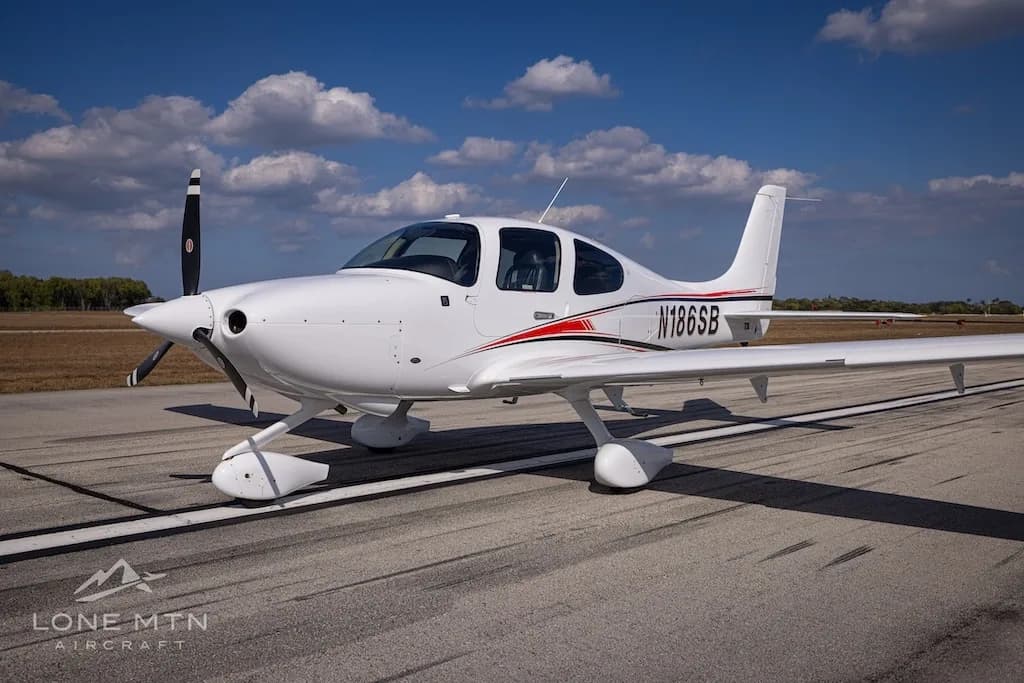 A white single-engine aircraft, N186SB, with red and black accent stripes, parked on an asphalt runway under a blue sky.