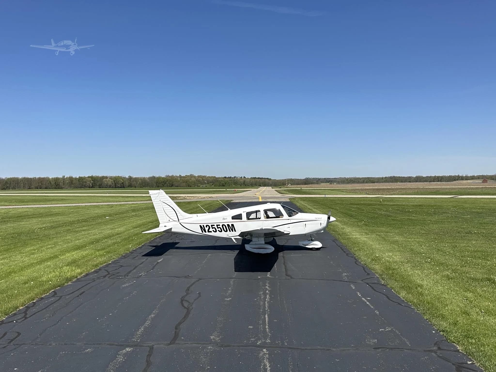 A white Piper Cherokee aircraft, N2550M, seen from a distance on an asphalt runway, surrounded by green fields.