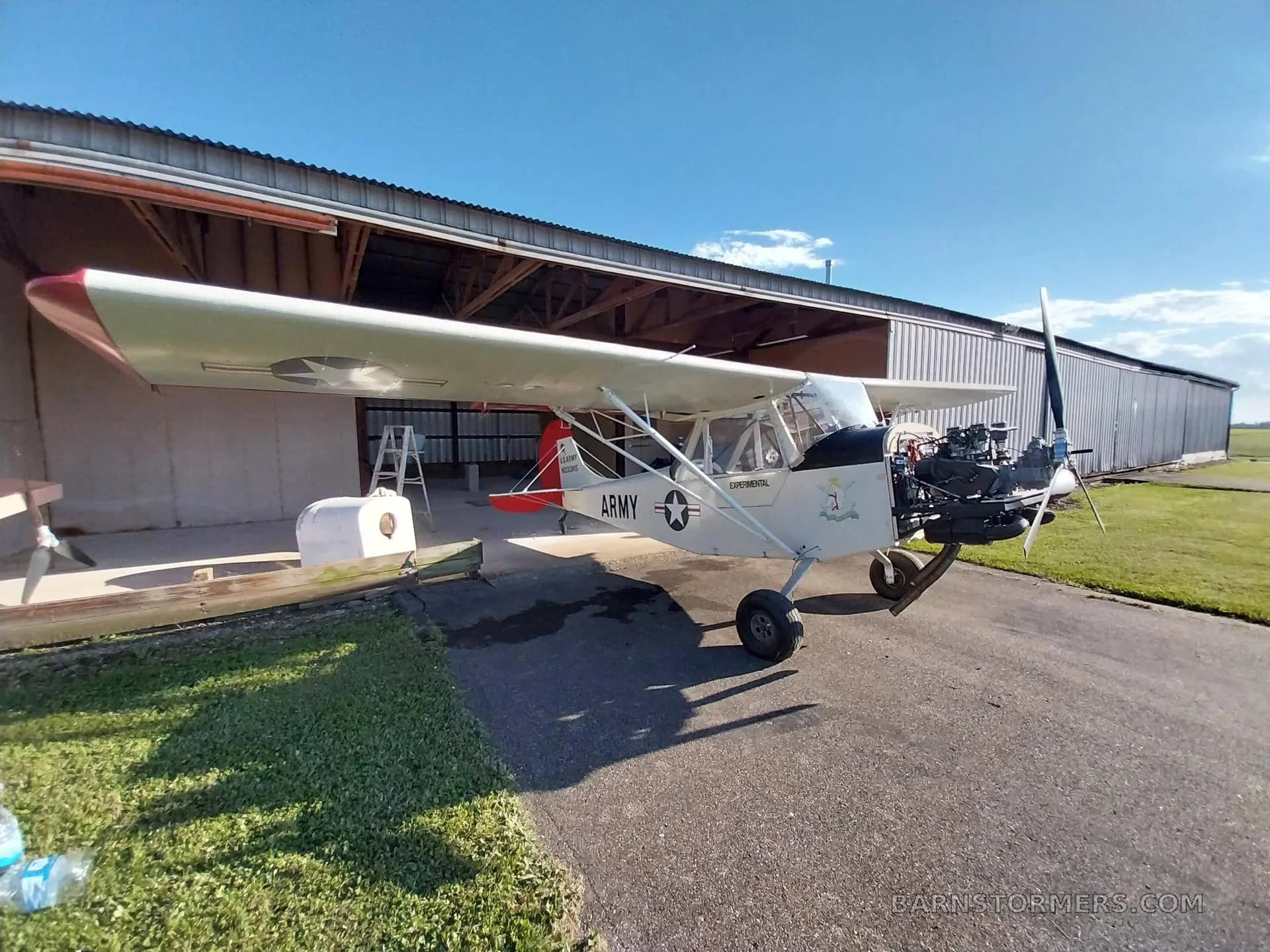A white experimental aircraft with 'ARMY' markings and an exposed engine sits on an asphalt ramp outside a hangar.