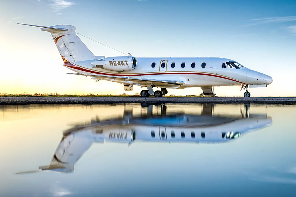 A white business jet with red and gold stripes (N24KT) is parked on a tarmac with its reflection in a puddle at sunset.