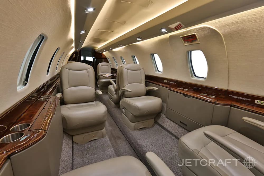The aircraft cabin shows beige leather seating arrangements and polished wood accents along the sidewalls.