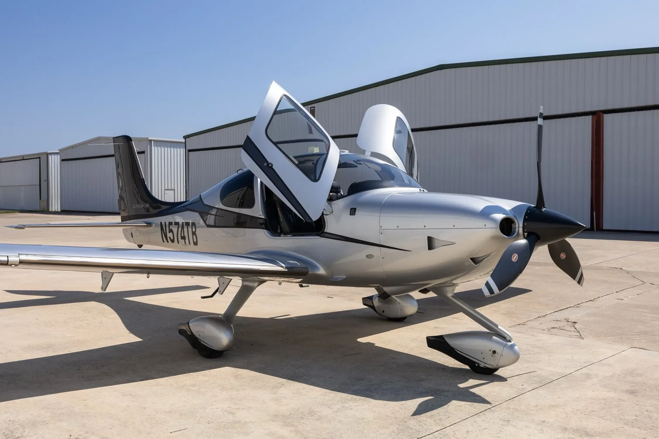A silver and black Cirrus SR22 aircraft, registration N574TB, parked on a tarmac with its gull-wing doors open.
