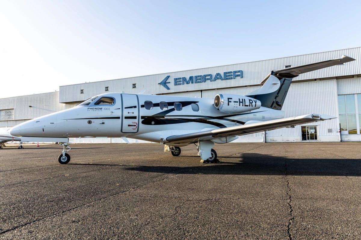 A white Embraer Phenom 100 business jet with black and white livery, registration F-HLRY, parked on an tarmac in front of an Embraer hangar.