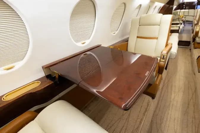 A close-up of a polished dark wood fold-out table and a cream leather seat next to a window in the aircraft cabin.