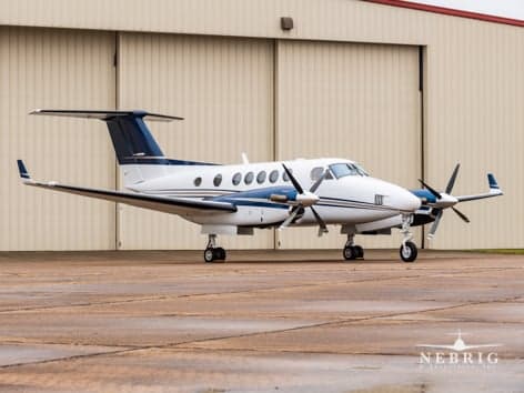A white and blue Beechcraft King Air 350 turboprop aircraft parked on a wet tarmac in front of a hangar.