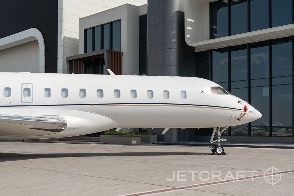 A close-up view of the front fuselage and nose of a white business jet with a gold and blue stripe, parked on an airfield.