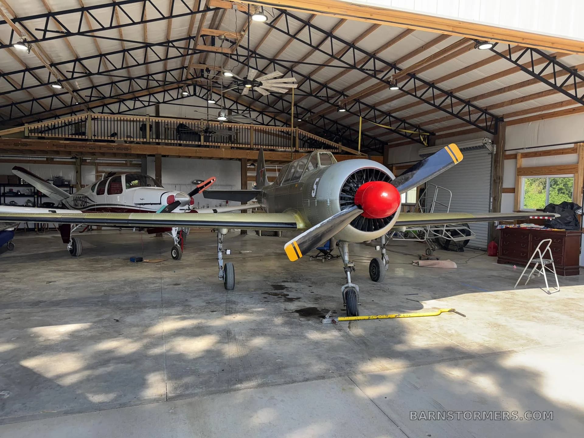 A grey single-engine propeller aircraft with a red spinner and yellow propeller tips, parked in a hangar next to a white and red twin-engine aircraft.