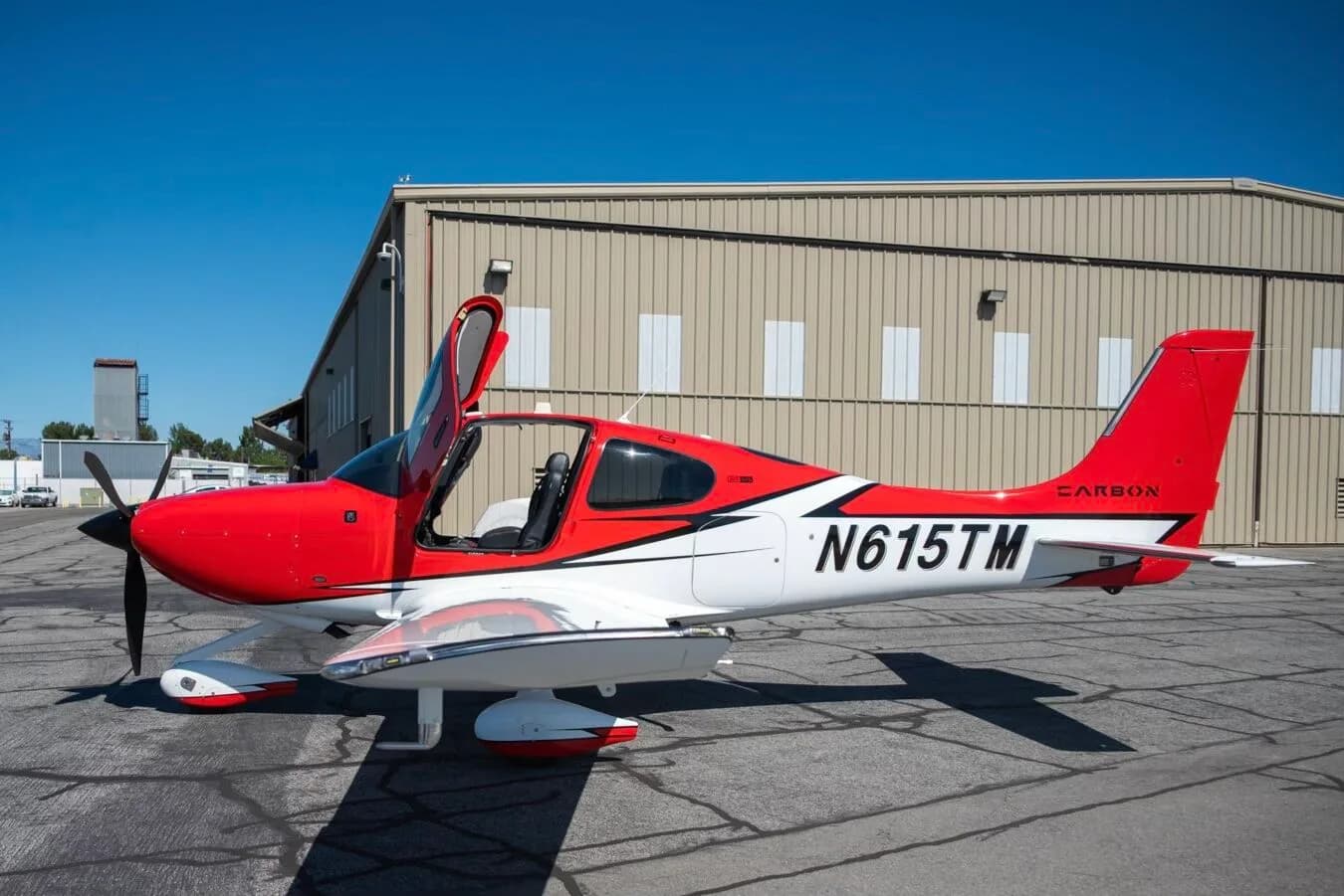 Side view of a red and white N615TM aircraft with its door open, parked in front of a hangar.