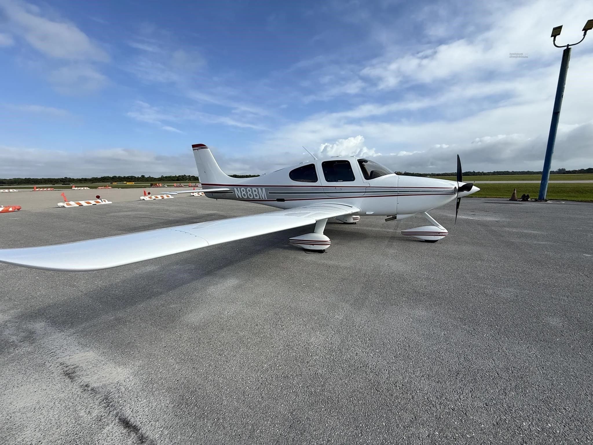 A white Cirrus SR22 aircraft with red and gray stripes, tail number N88BM, parked on an airport tarmac, showing the left side.