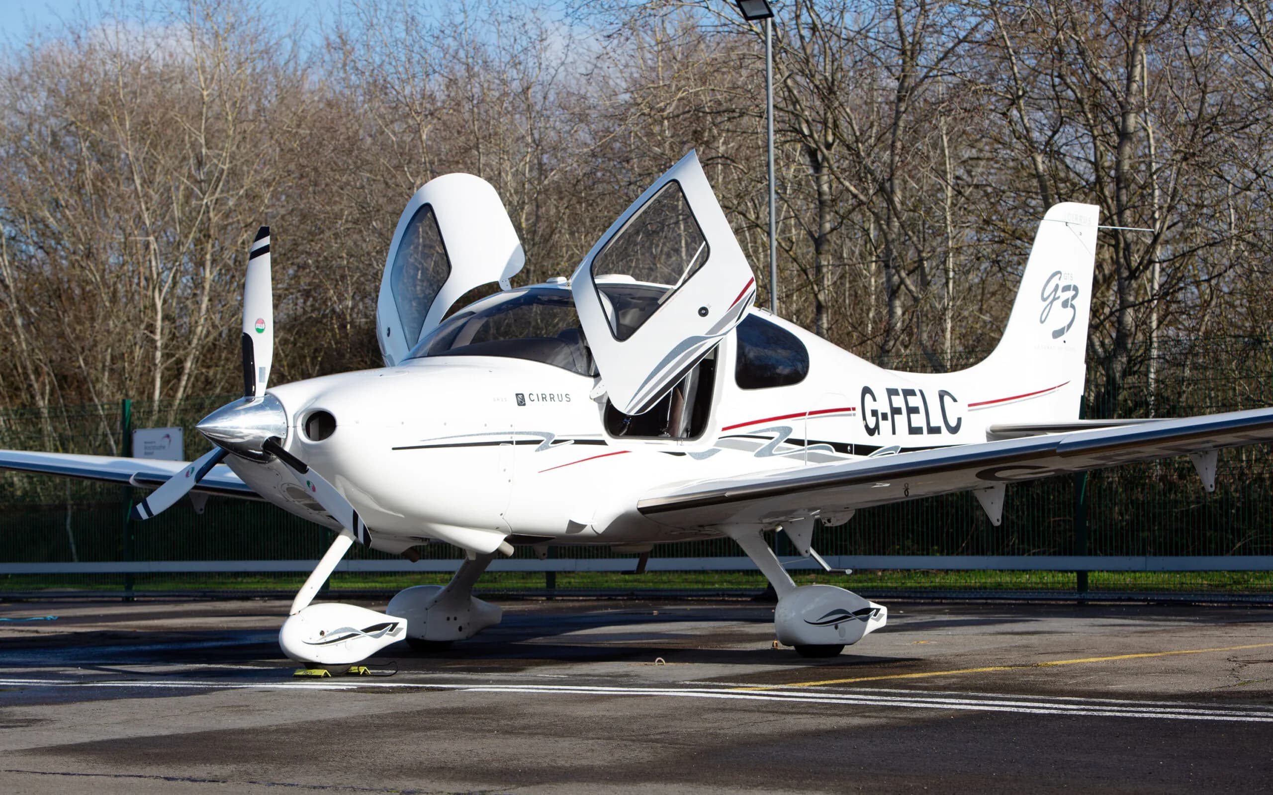 A white Cirrus SR22 G3 aircraft with red and black stripes, registration G-FELC, parked on a tarmac with its gull-wing doors open, viewed from the front-right.