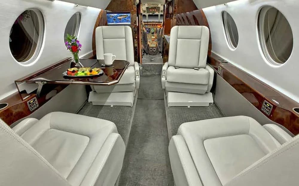 A view of the business jet cabin interior showing light gray seats, a dining table with a meal, and flowers.