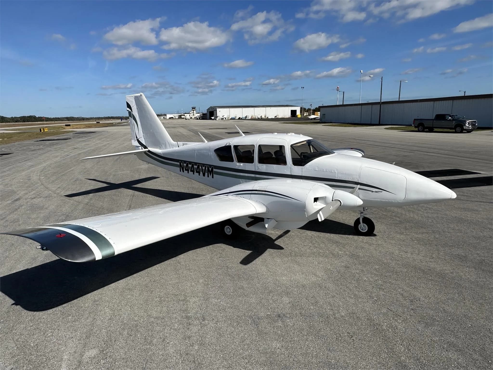 A three-quarter rear view of a white Piper aircraft with green and black stripes, registration N444VM, parked on an airfield.