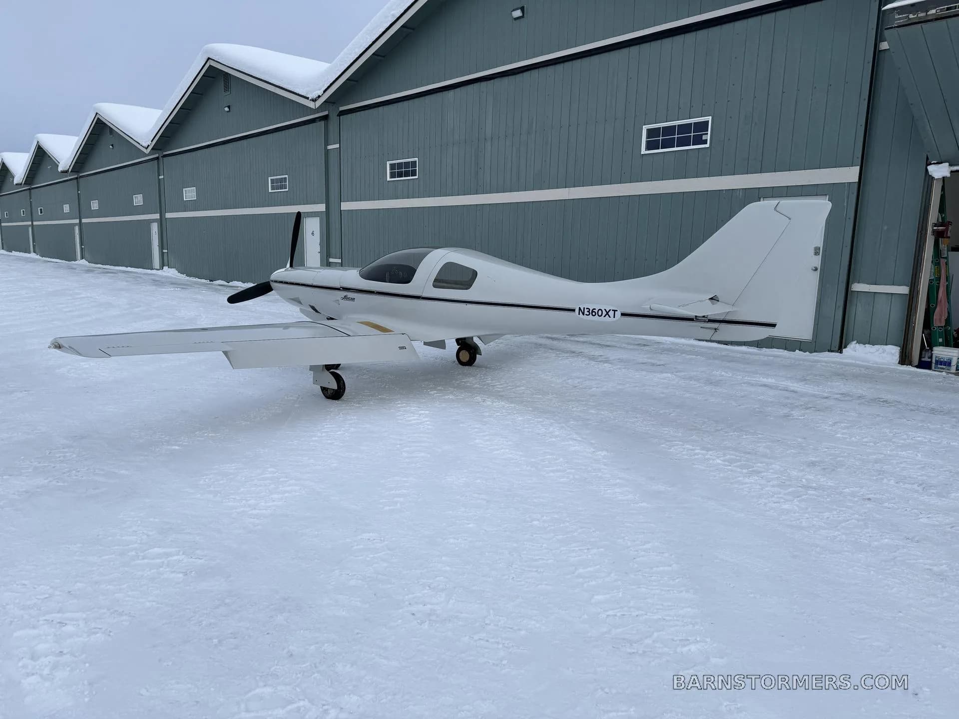 A white Lancair Legacy aircraft (N360XT) with a black stripe is parked on snowy ground in front of a row of blue hangars.