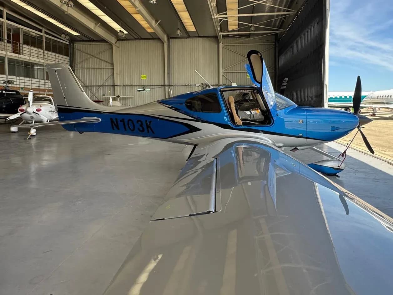 A blue and silver Cirrus SR22 aircraft (N103K) with open doors, viewed from the left side in a hangar.