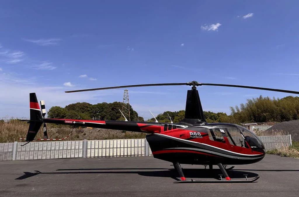 A black and red Robinson R66 Turbine helicopter is parked on an asphalt surface with trees and a fence in the background.