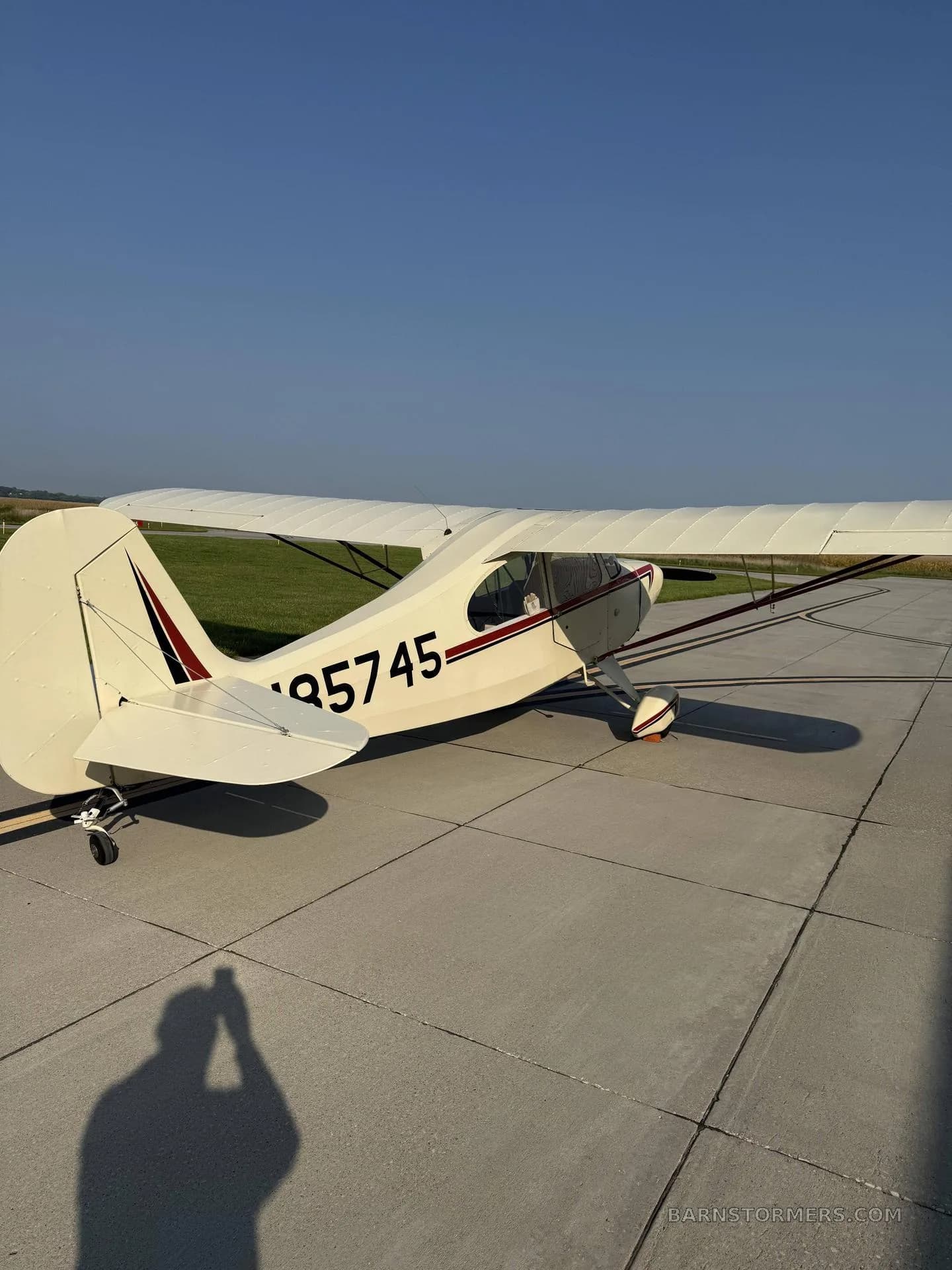 A cream-colored, high-wing taildragger aircraft with red and black stripes, registration N85745, viewed from the rear on a concrete tarmac.