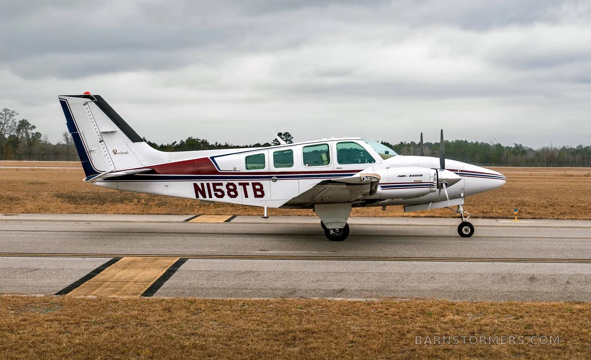 A white twin-engine Beechcraft Baron N158TB with maroon and blue stripes is seen from the side on a taxiway under a cloudy sky.