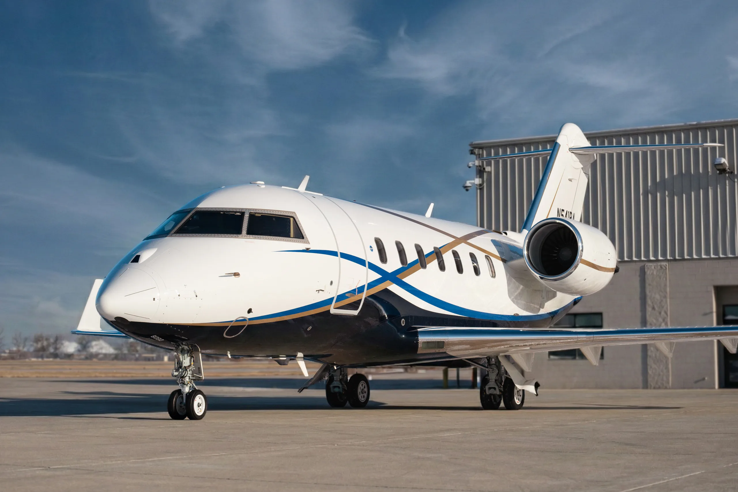 A white and blue Bombardier Challenger 350 business jet, registration N541BA, is parked on a tarmac in front of a hangar under a partly cloudy sky.