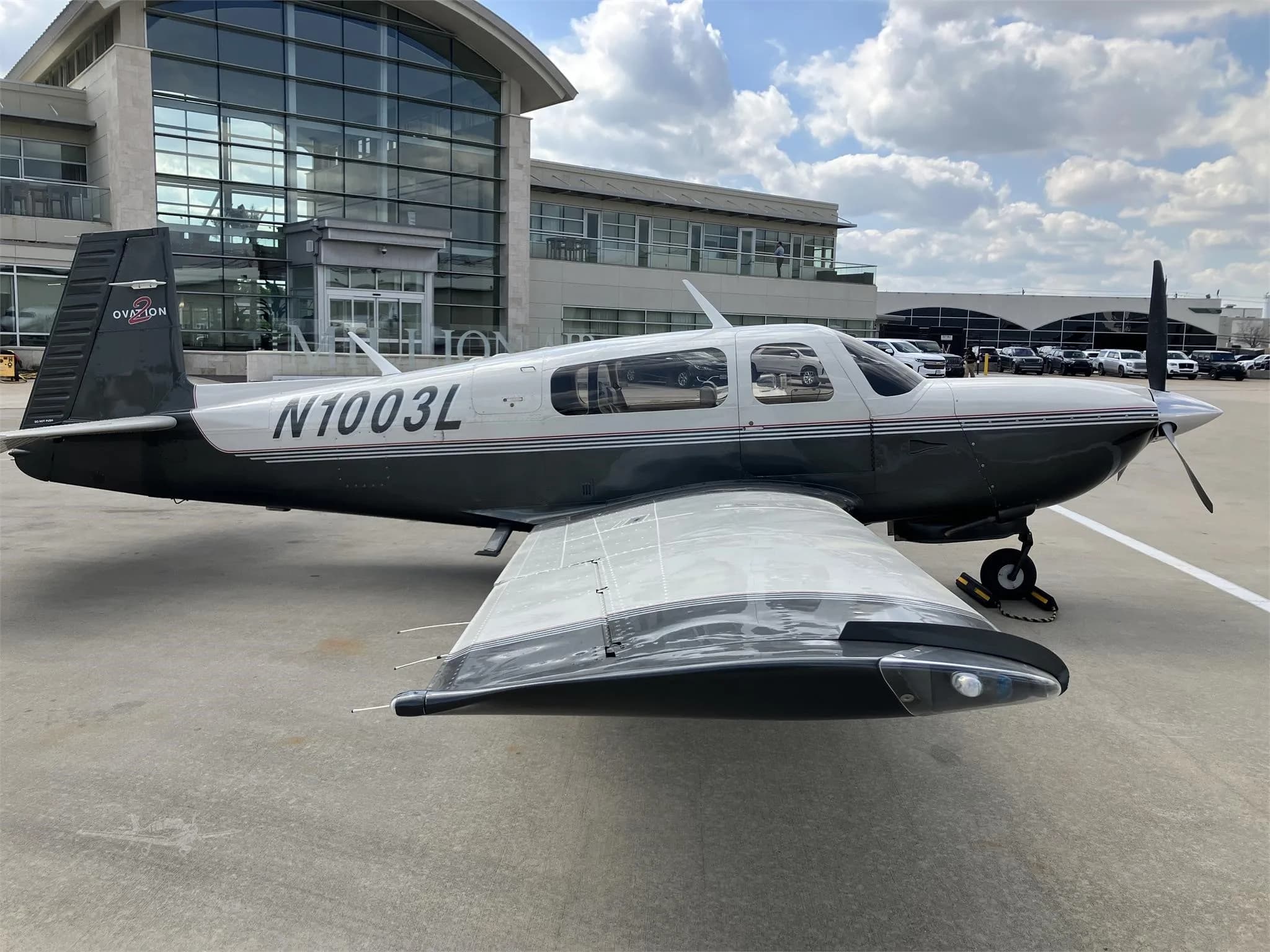 A dark grey and white Mooney M20 Ovation aircraft with registration N1003L is parked on an airport apron in front of a modern building.