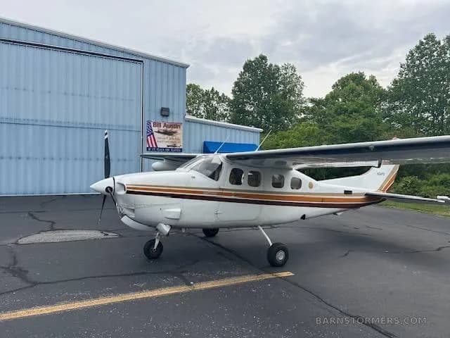A white Cessna 337 Skymaster with brown and orange stripes is parked on an asphalt ramp in front of a blue hangar.