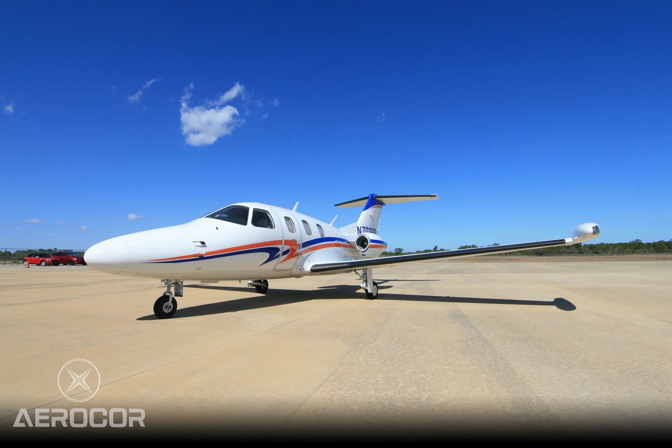 A white Eclipse 500 jet with blue and orange stripes, tail number N703BR, parked on an airfield under a clear blue sky.