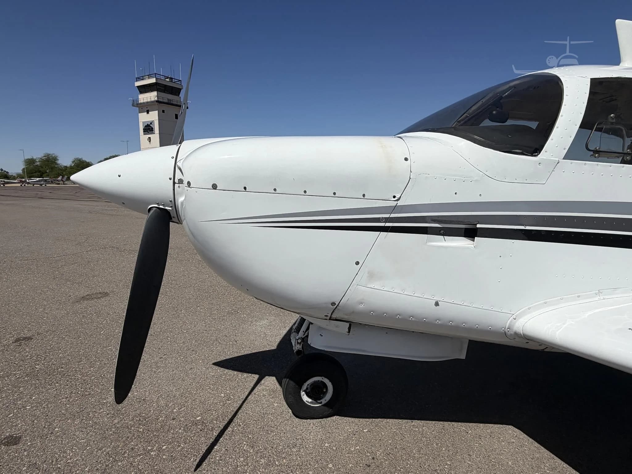 Close-up of the nose and propeller of the Mooney M20J Super 21SE aircraft with a control tower in the background.