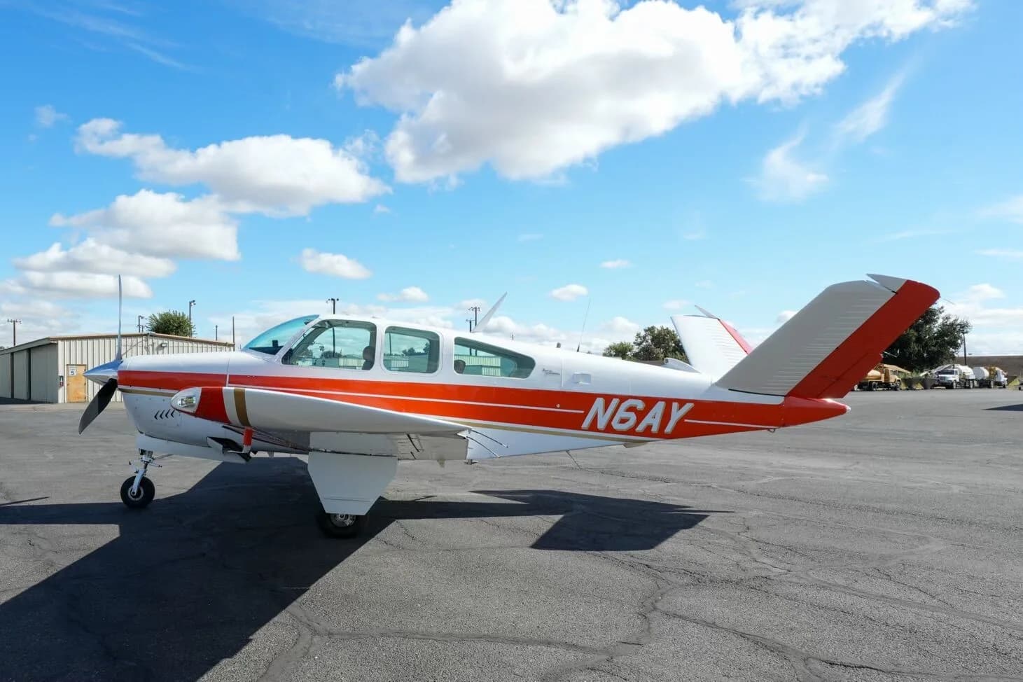 A white and orange Beechcraft Bonanza N6AY parked on an airport tarmac with hangars in the distance.