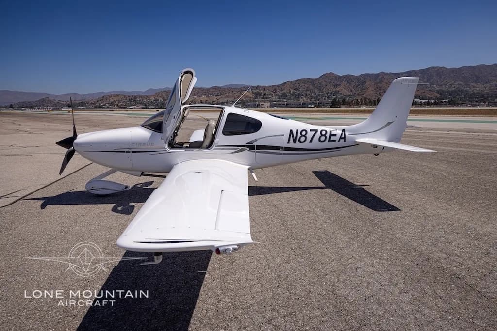 A white Cirrus aircraft with registration N878EA and open doors is parked on an airfield, viewed from the rear-right.