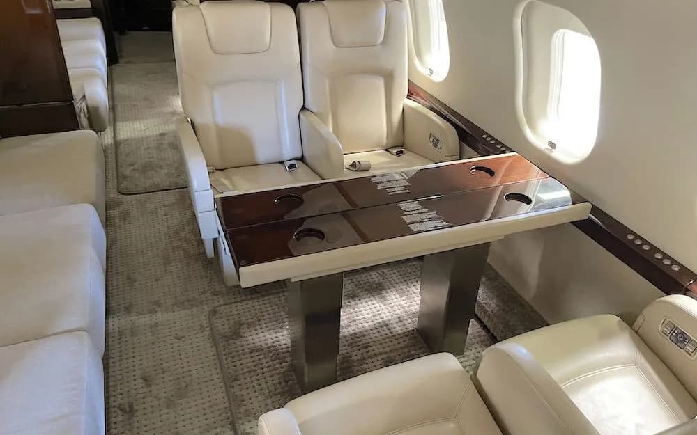 A private jet cabin showing two cream-colored seats facing a polished wooden table with cup holders.