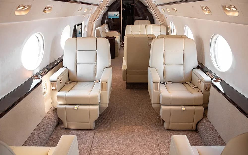 A wide view of a luxurious aircraft cabin with rows of beige leather seats and large oval windows.